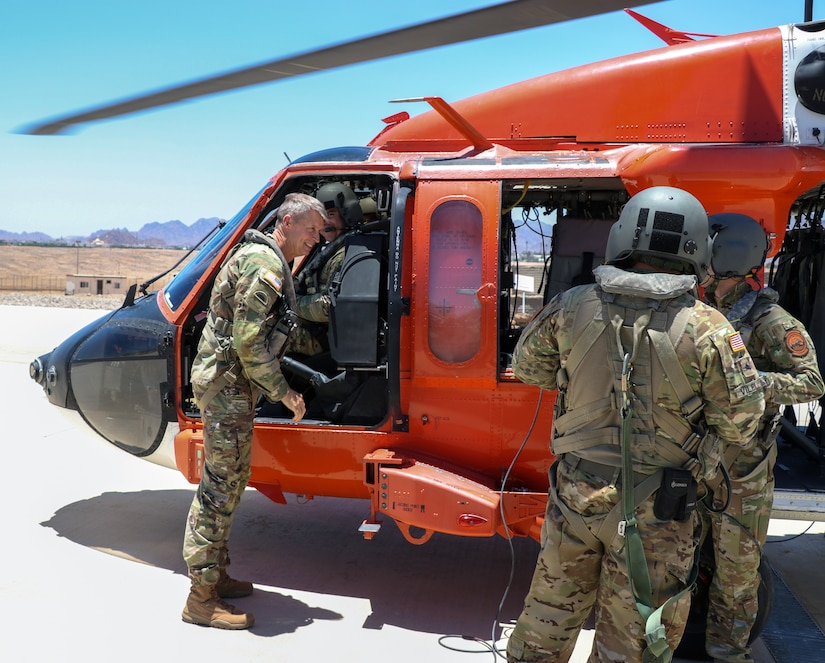 Service members stand by an orange helicopter on a flightline.
