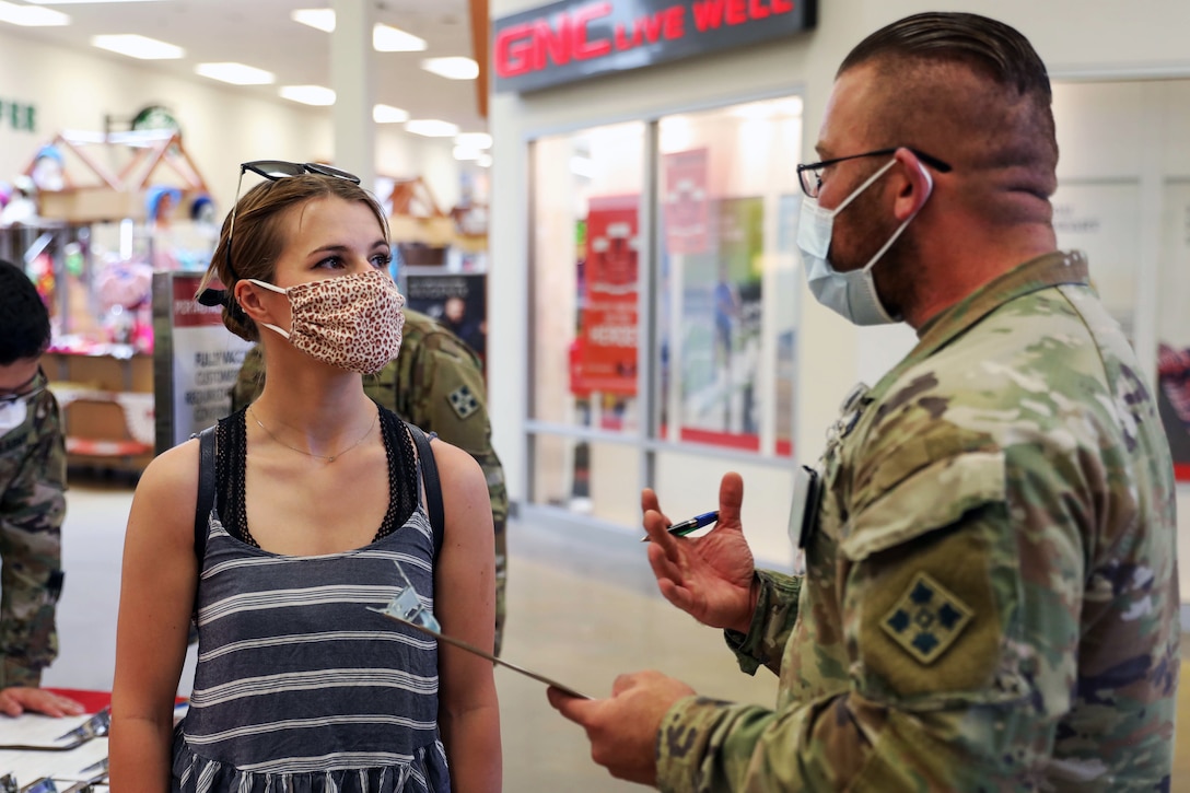 A soldier wearing a face mask and holding a clipboard explains to a woman wearing a face mask how to get a COVID-19 vaccine.