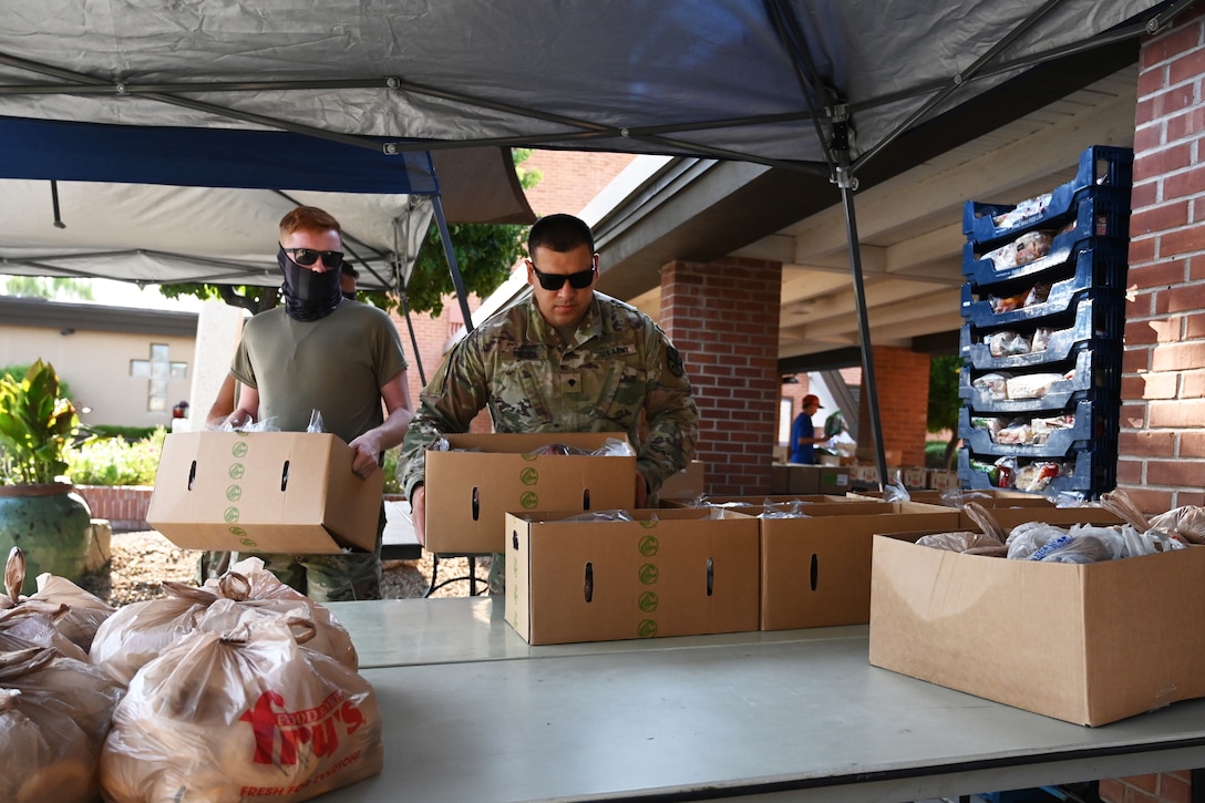 Two guardsmen, one wearing a face mask, carry boxes of prepared groceries to be donated to area residents.