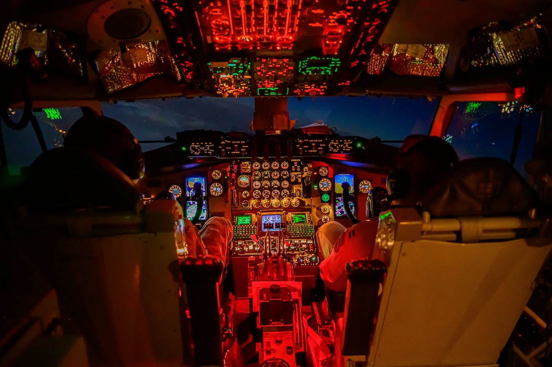 Airmen, shown from behind, sit in a cockpit illuminated with different colored lights.