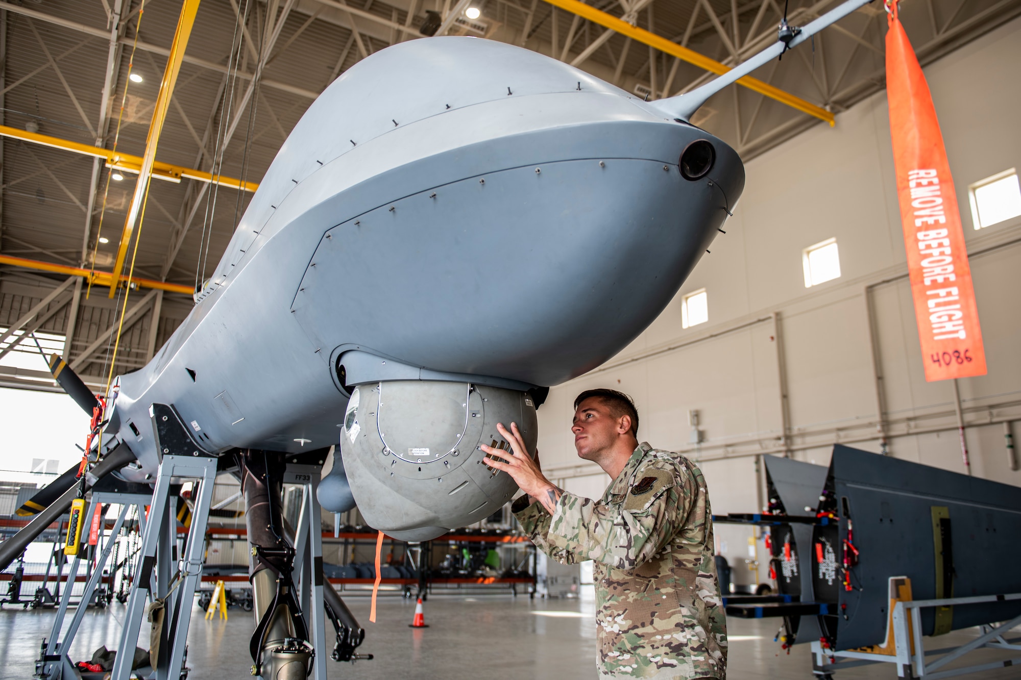 Senior Airman Levi stands under an MQ-9 Reaper and inspects the multi-spectral targeting system for flaws.