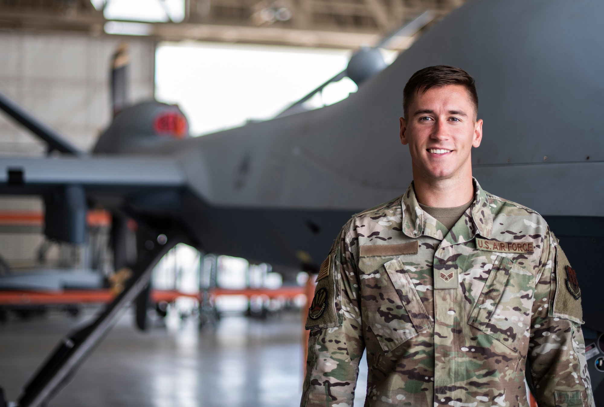 Senior Airman Levi stands to the side of an MQ-9 Reaper and smiles for a photo.