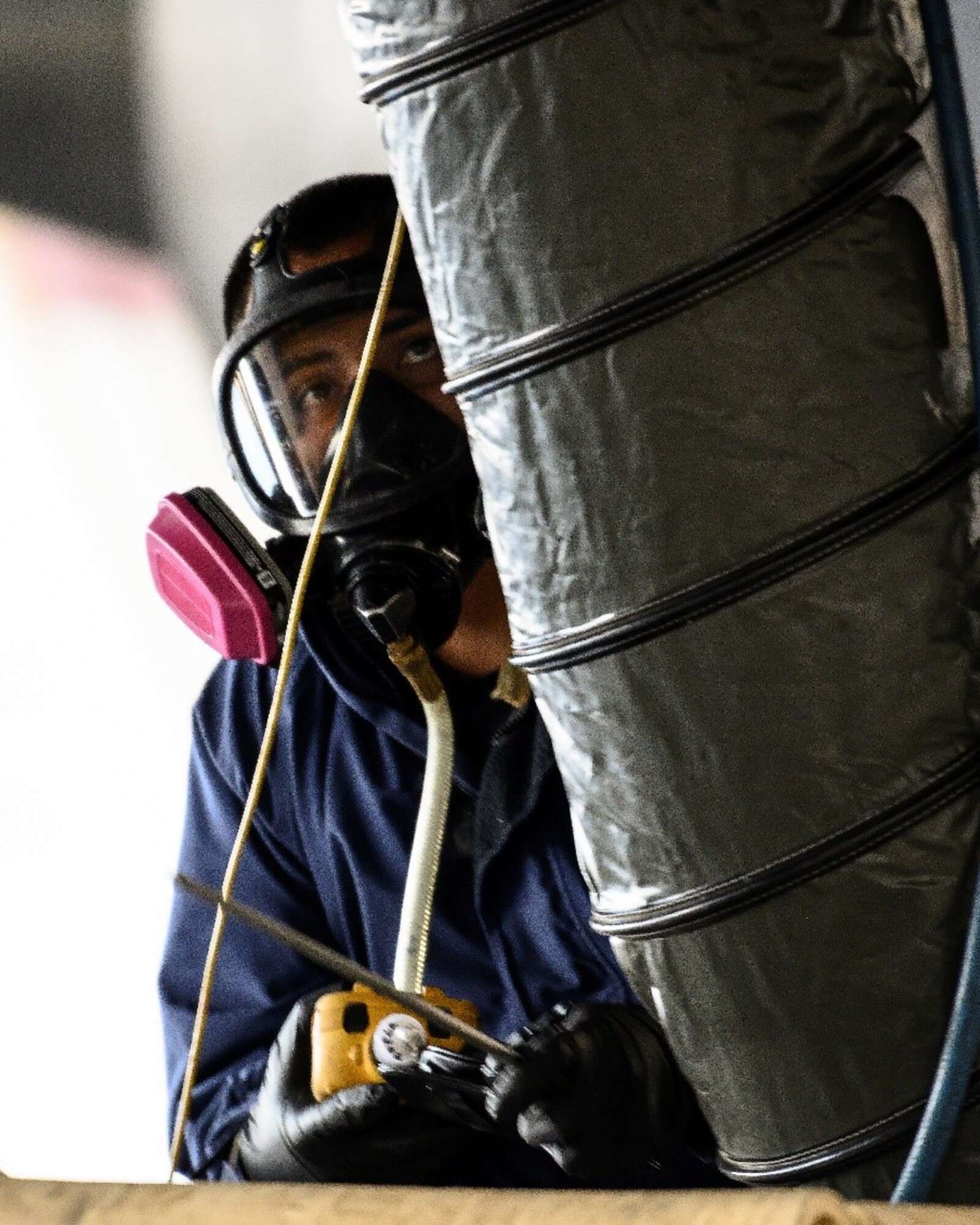 An Airman in a respirator looks up at a long tube that's feeding into the compartment of an aircraft.
