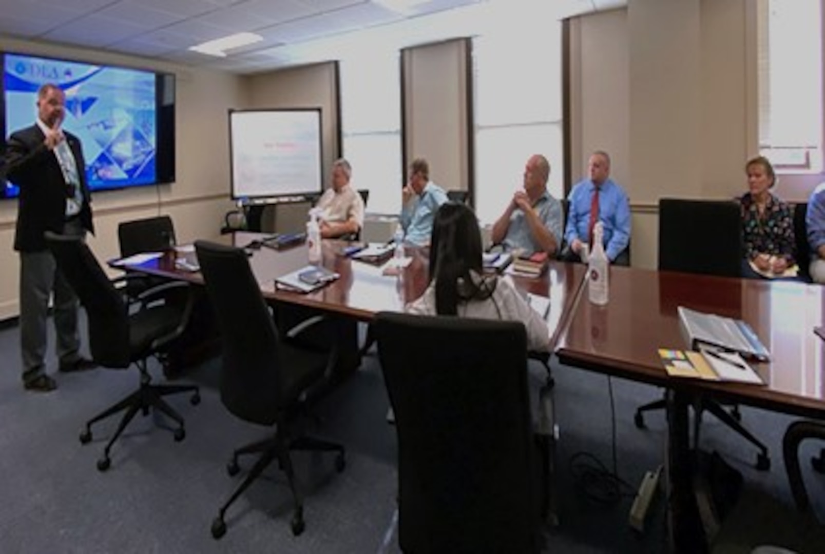 Man stands in front of conference room table presenting to seated participants