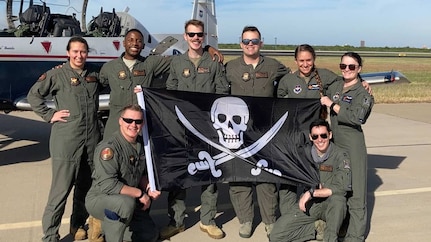 Eight pilots pose holding black "FAIP" flag with skull and swords