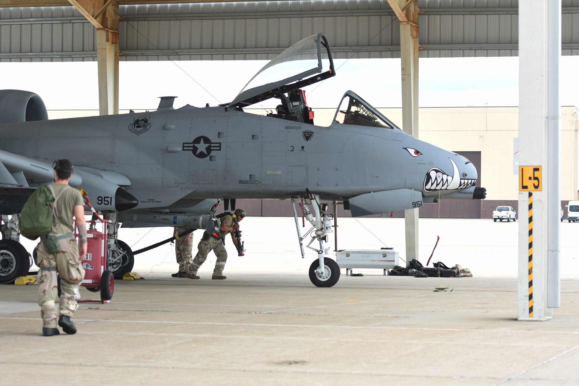 An Airman in chemical protective gear walks toward an A-10 Thunderbolt II, while another Airman drags a fuel hose toward it.