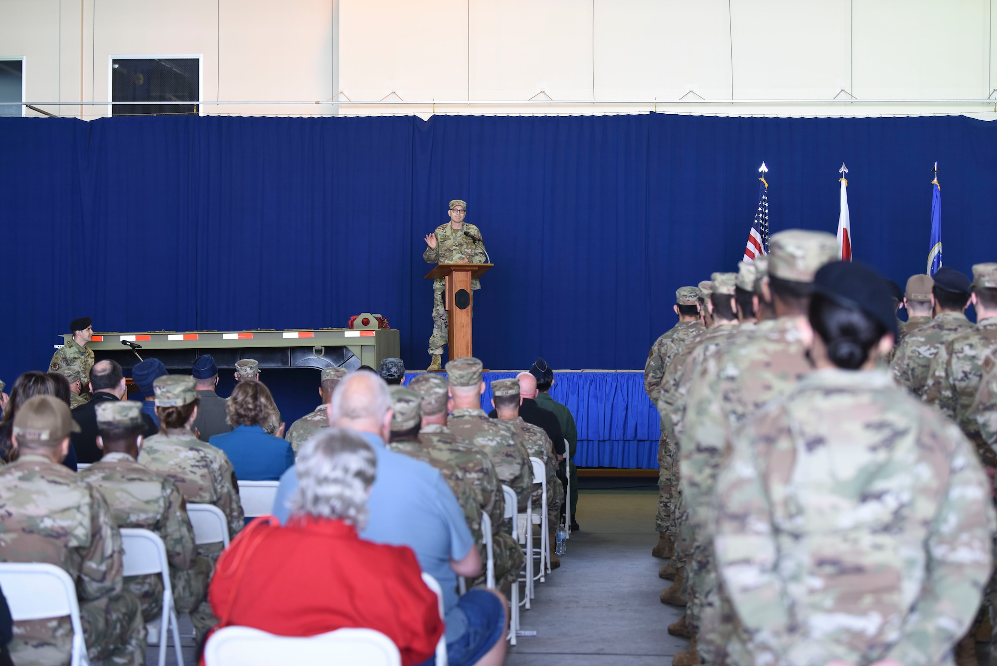 A military member in uniform stands on a stage behind a podium speaking to a crowd of civilians and military members seated to the left side and a flight of uniformed military members standing at parade rest at the right side.