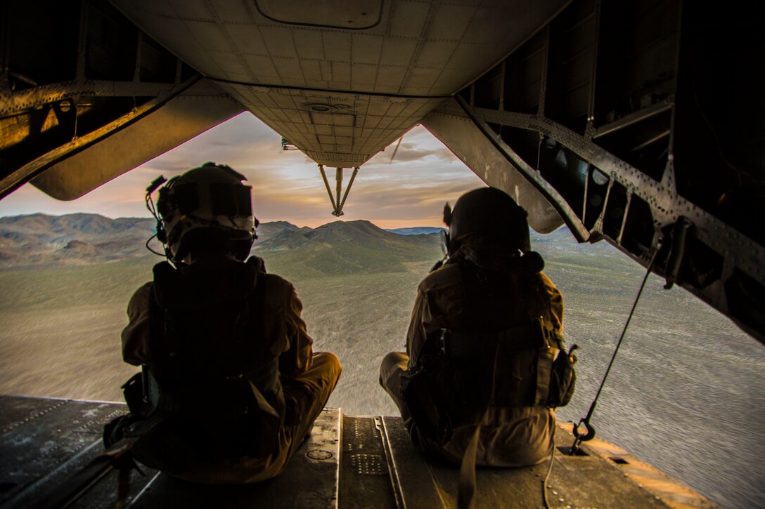Two Marines look out over barren terrain from an aircraft in flight.