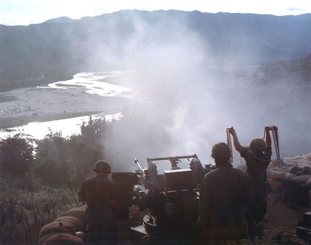 A soldier holds a string of ammunition above his head as two other soldiers work to shoot a large machine gun into a valley.