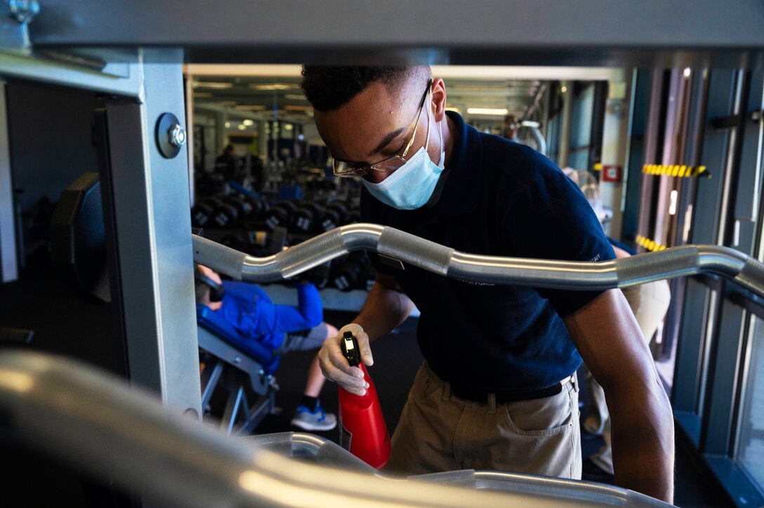 An airman wearing a face mask and gloves holds a bottle of sanitizer as he wipes down gym equipment.