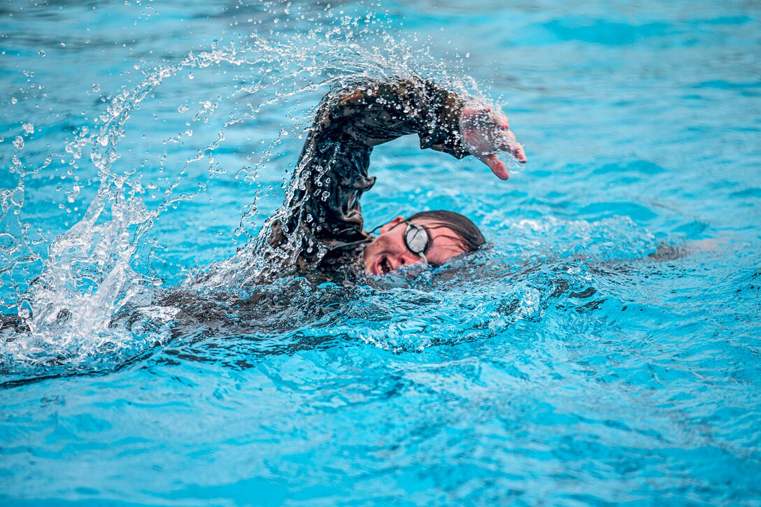 A Marine swims in a pool while wearing goggles and a full uniform.