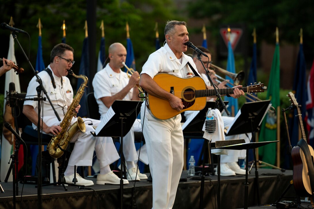 A sailor sings while playing a guitar with a band on stage.