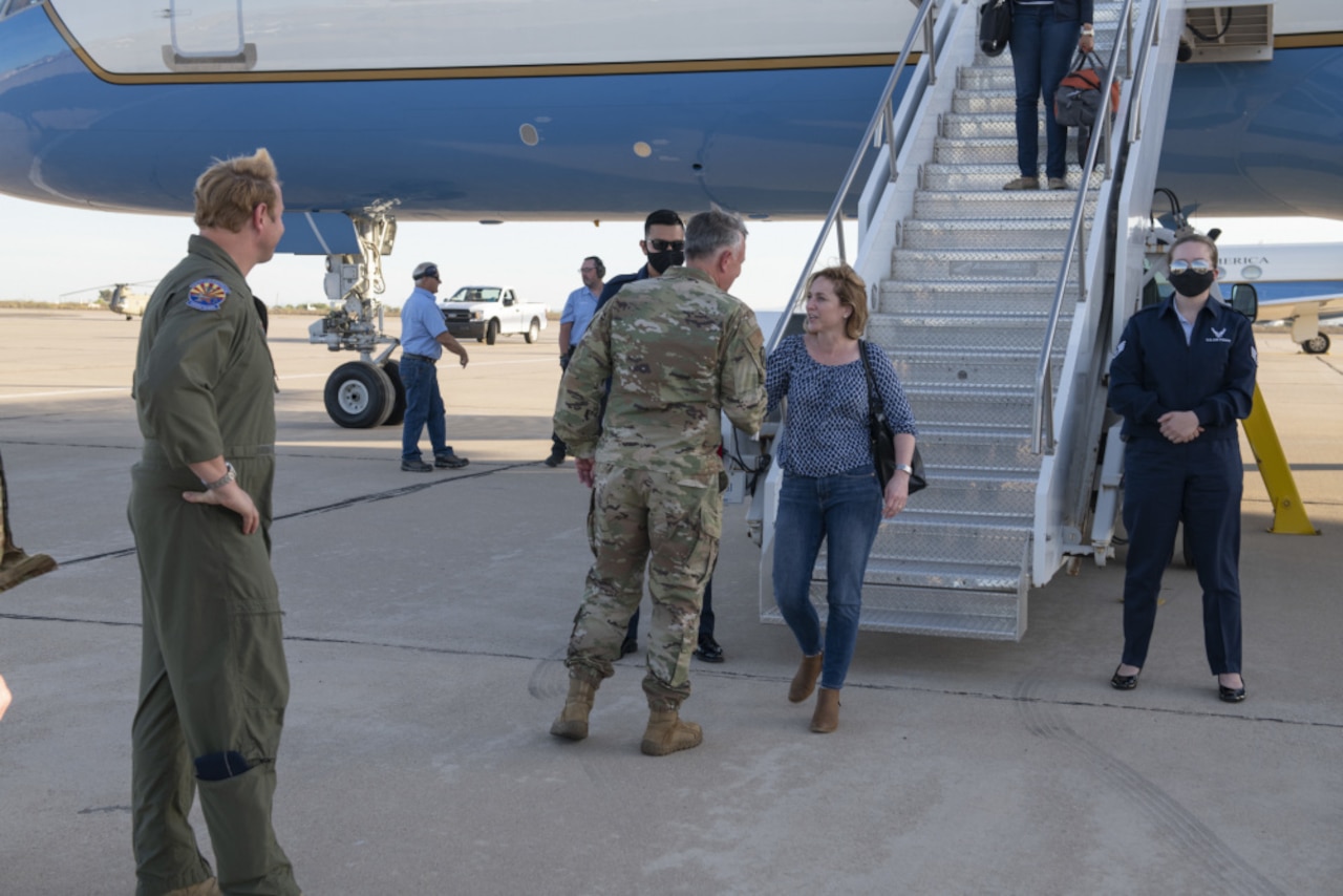 On a tarmac, a man in a military uniform shakes the hand of a woman who has descended the steps of an airplane.