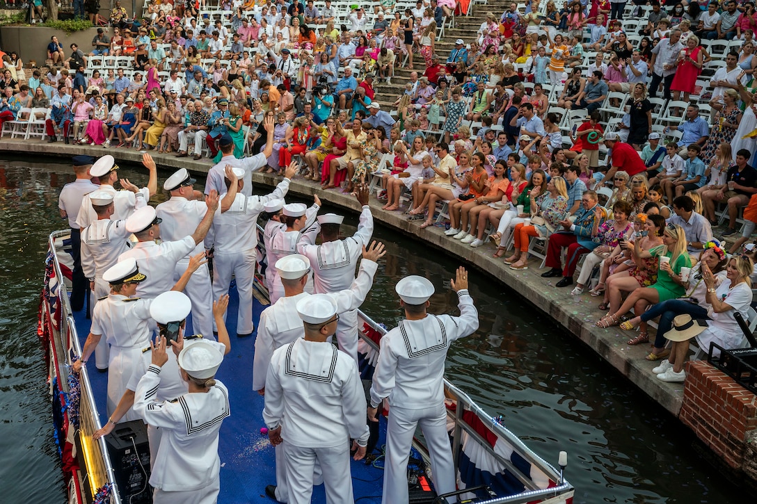 A group of service members wave to a crowd from a boat on the water.