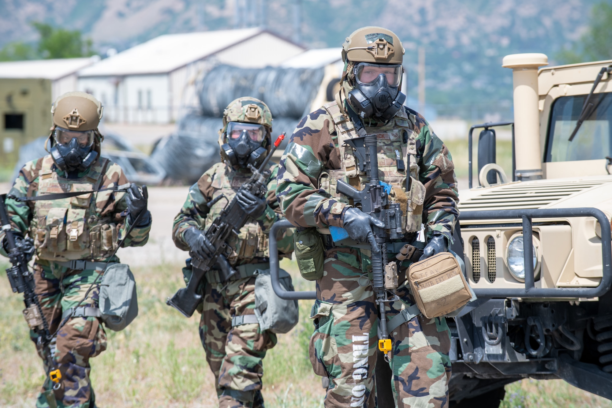 Three Airmen holding rifles and in chemical gear walking in front of a military Humvee.