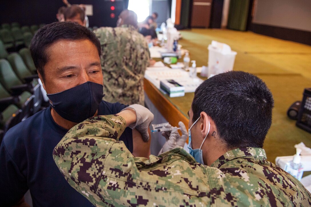 A sailor wearing a face mask and gloves gives an injection into the arm of a man.