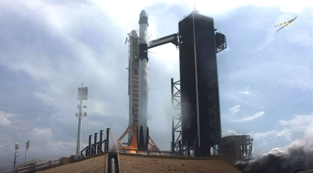 A rocket sits on a launch site, as flames and smoke billow from beneath.