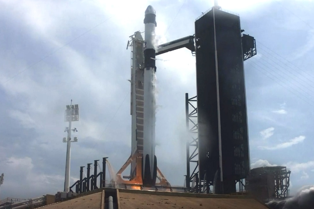 A rocket sits on a launch site, as flames and smoke billow from beneath.