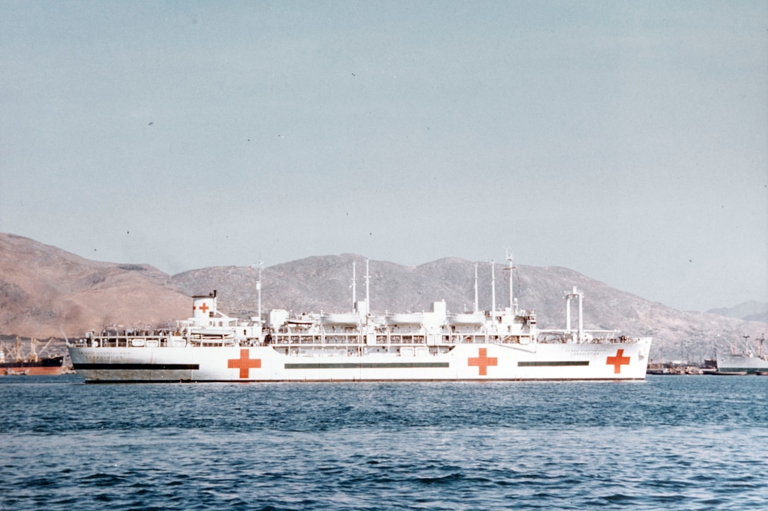 A white Navy ship with red crosses painted on its hull travels in blue water.