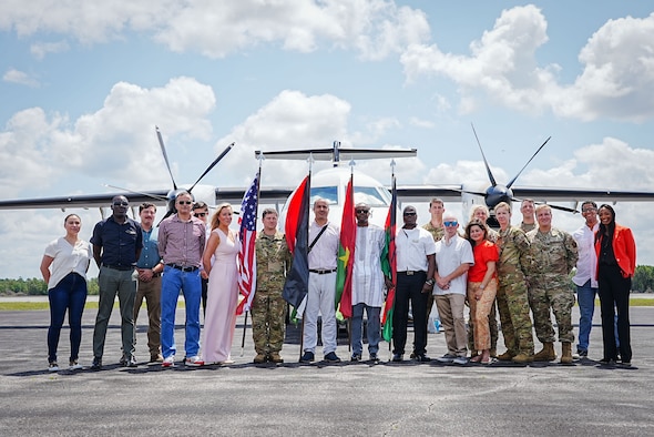 Group of Airmen, civilians and students pose for a photo in front of an aircraft while holding flags.