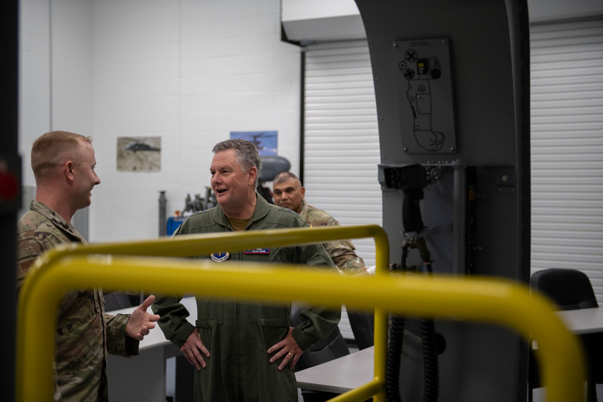A photo of two Airmen standing behind a yellow bar attached to an HH-60 simulator.