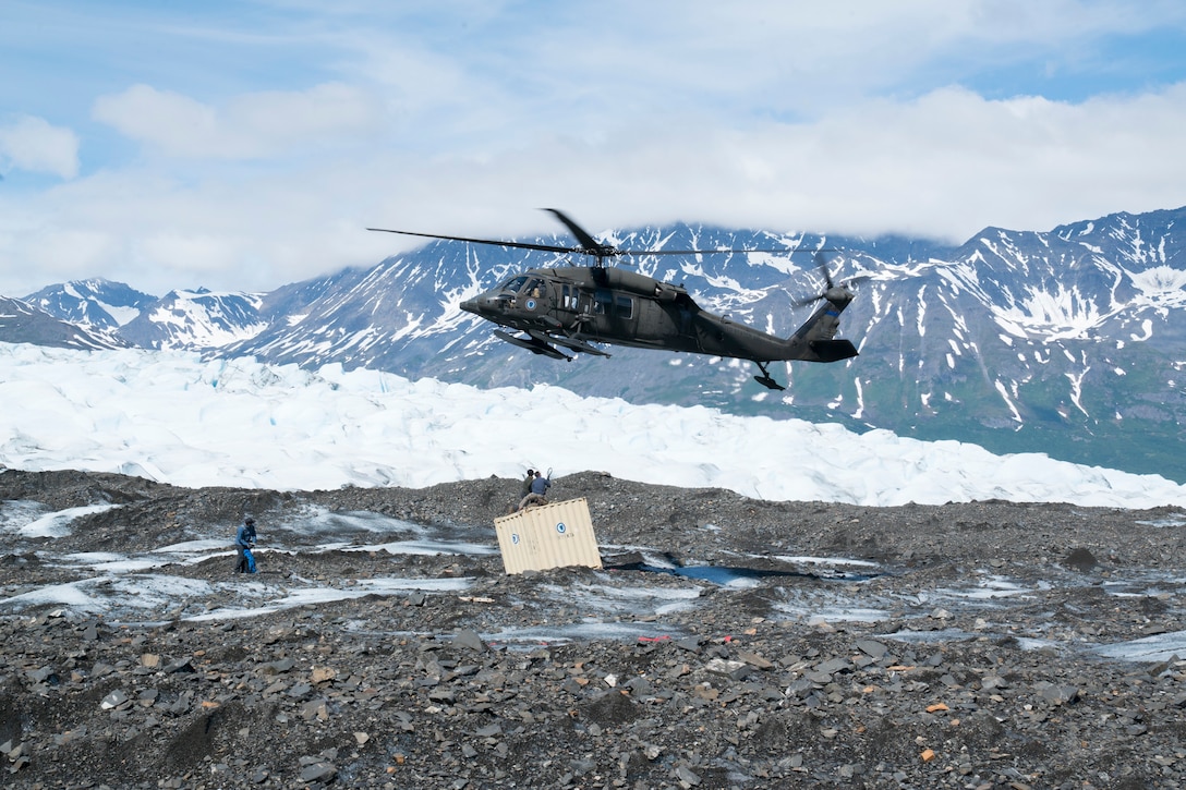 A helicopter flies just above the ground with a cable attached to a container in a mountainous area.