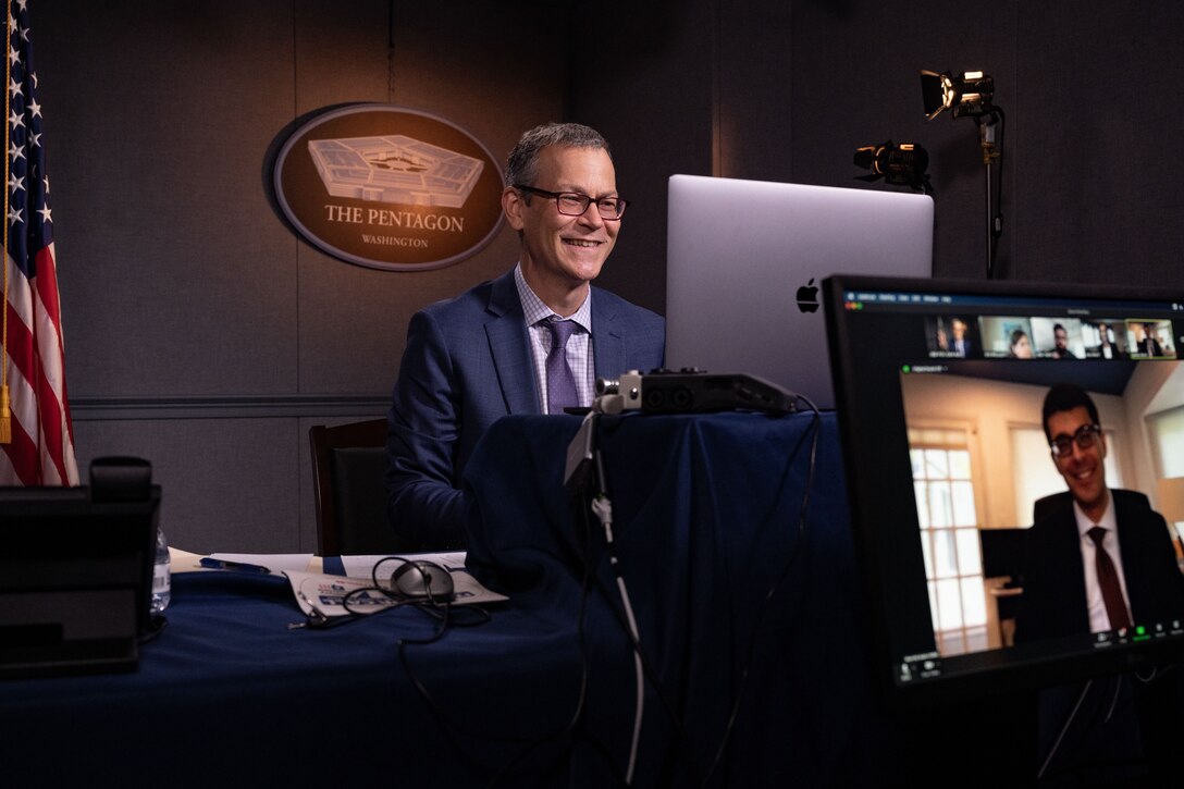 A man sits in a studio behind a laptop. Nearby, a monitor displays the face of another man.