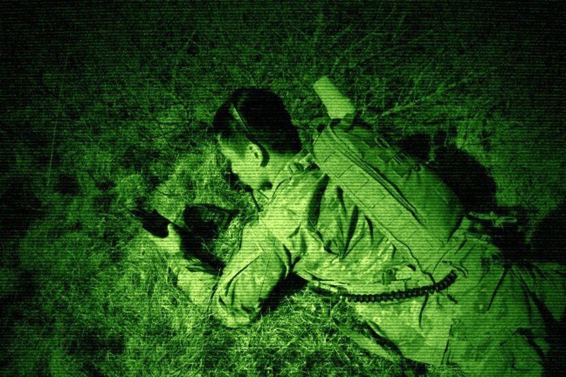 A soldier lays on the ground holding a communication device, illuminated by green light.