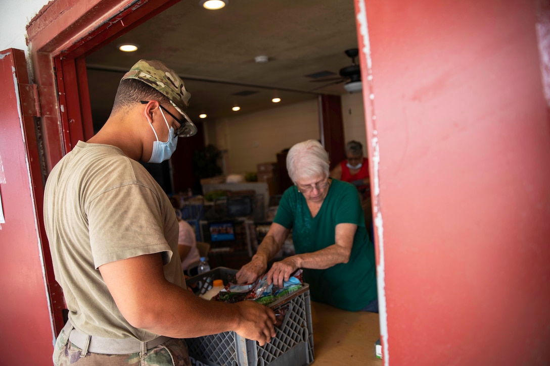 A soldier wearing a face mask gives a crate of food to an elderly woman.