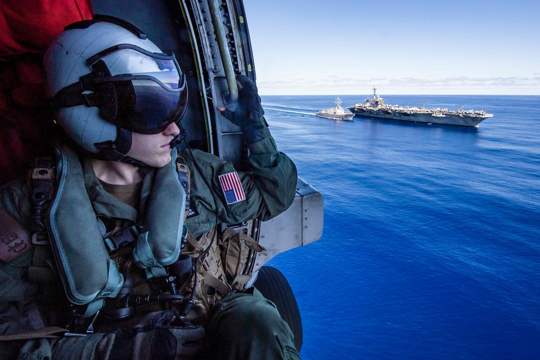 A sailor sits inside a helicopter while looking out toward two ships traveling through waters.