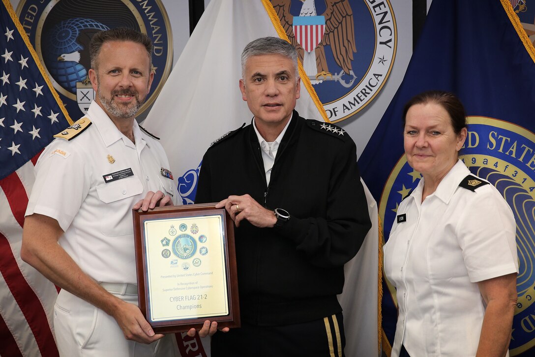 Royal Canadian Navy Commodore Matthew Bowen (left), J5 Multinational Partnership and Integration vice, accepts a plaque from U.S. Army Gen. Paul M. Nakasone, U.S. Cyber Command commander and National Security Agency director, and Command Sgt. Major Sheryl D. Lyon, USCYBEERCOM and NSA senior enlisted leader, on behalf of Team 15, the champions of Cyber Flag 21-2, June 25, 2021. The exercise allowed cyber operators to train in a tough competitive environment and identify characteristics of successful cyber protection teams, enabling USCYBERCOM to proactively develop successful teams at scale.