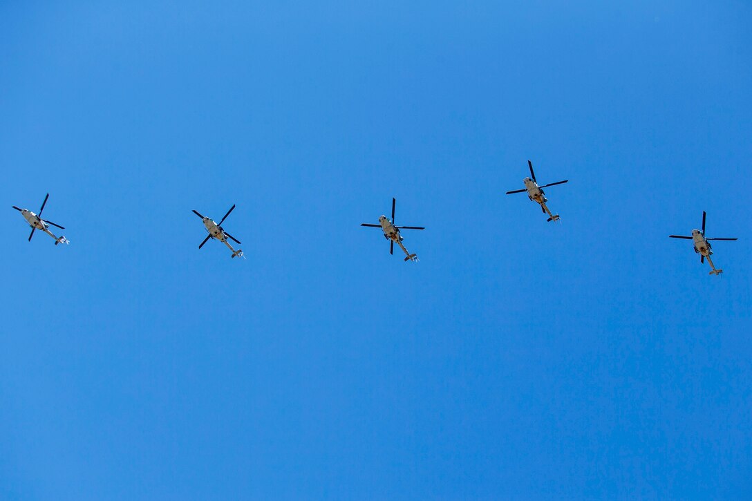 Five Marine Corps helicopters, viewed from below, fly in a line in blue sky.