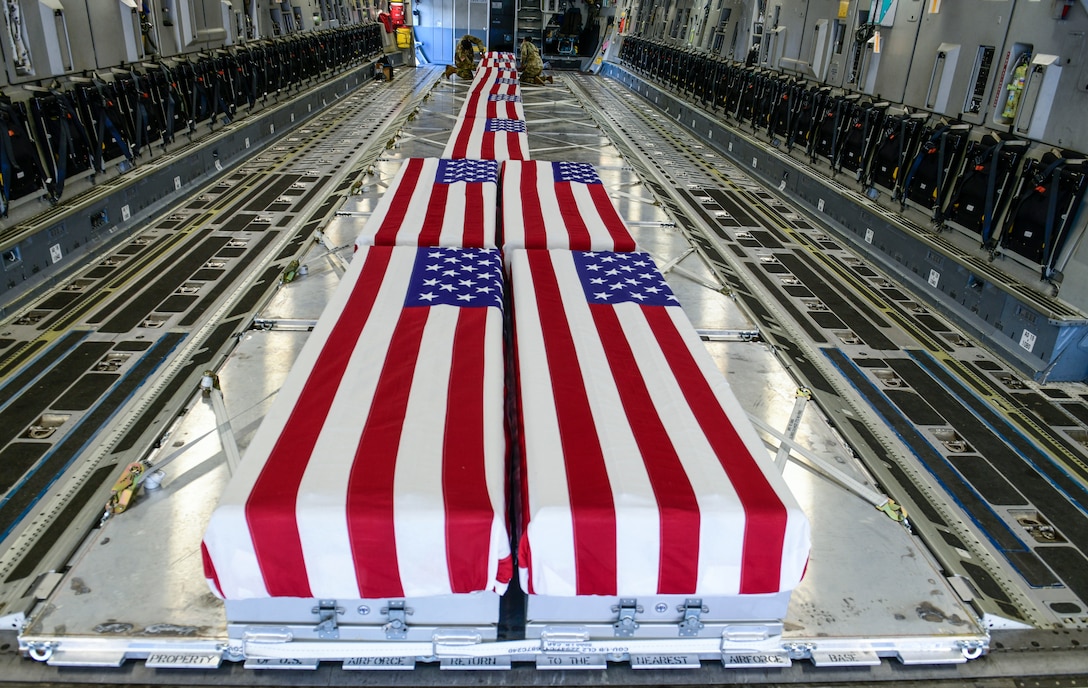 At least 10 large cases are draped with U.S. flags. In the background, two service members attach a flag to a case.