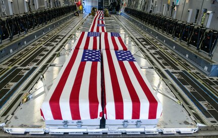 At least 10 large cases are draped with U.S. flags. In the background, two service members attach a flag to a case.
