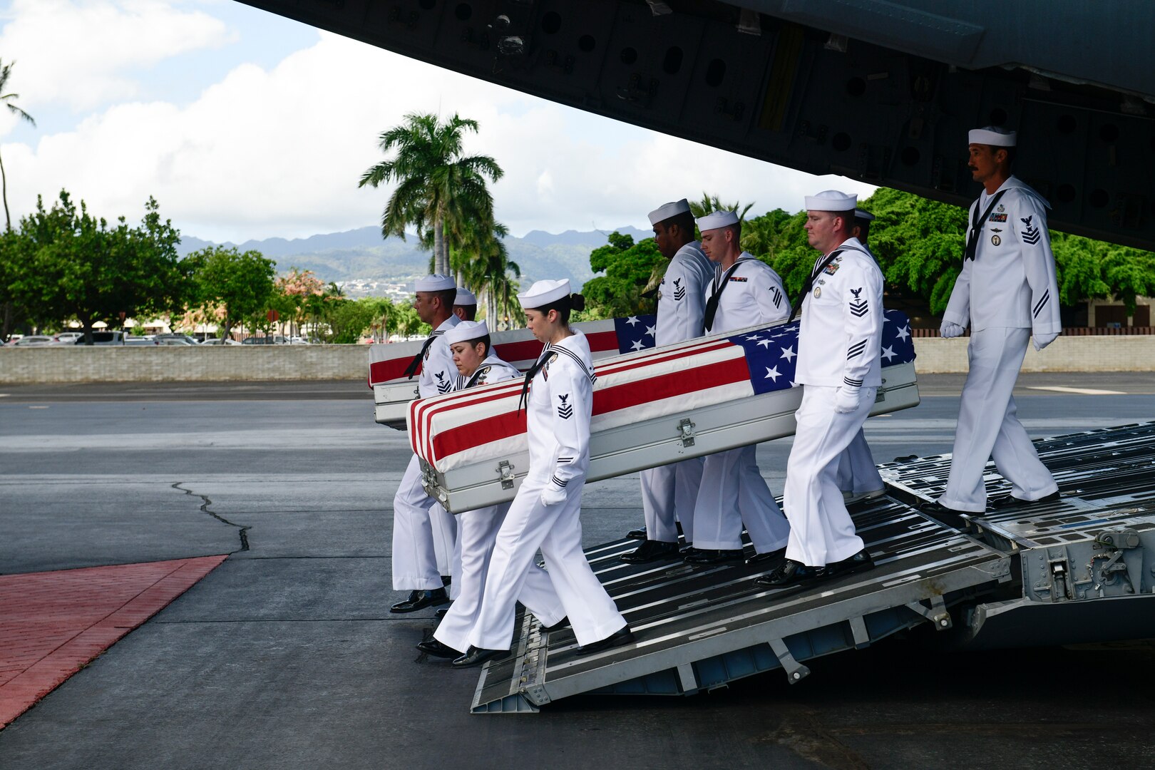 Sailors carry two flag-draped caskets down the cargo ramp of an airplane.