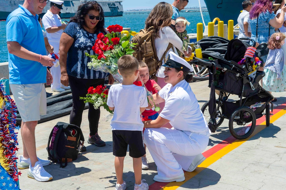 A sailor kneels next two children holding flowers as other stand around.