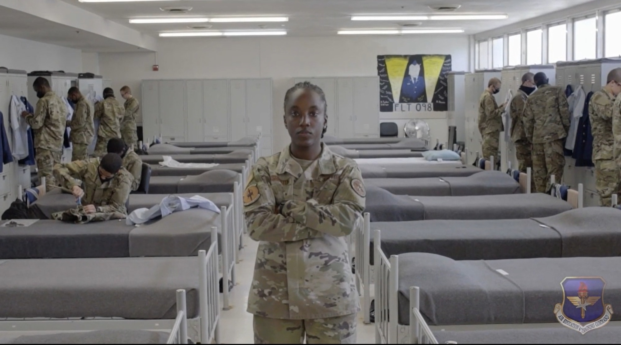 Staff Sgt. Alexandria Washington stands with her arms crossed while trainees are at their lockers in the background.