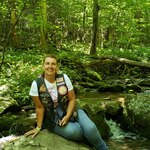 Women in a white shirt and brown vest leans against a rock in the woods