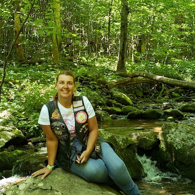 Women in a white shirt and brown vest leans against a rock in the woods