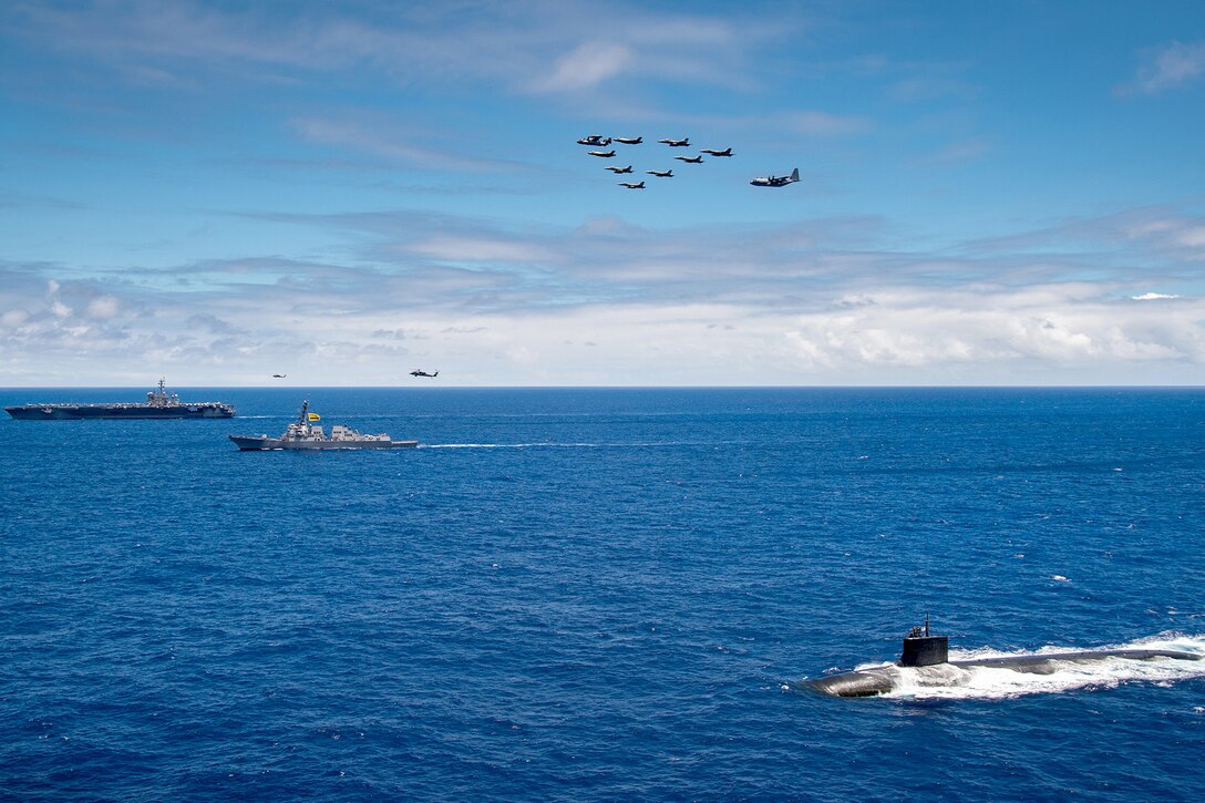 Two ships and a submarine sail in formation while aircraft fly above.