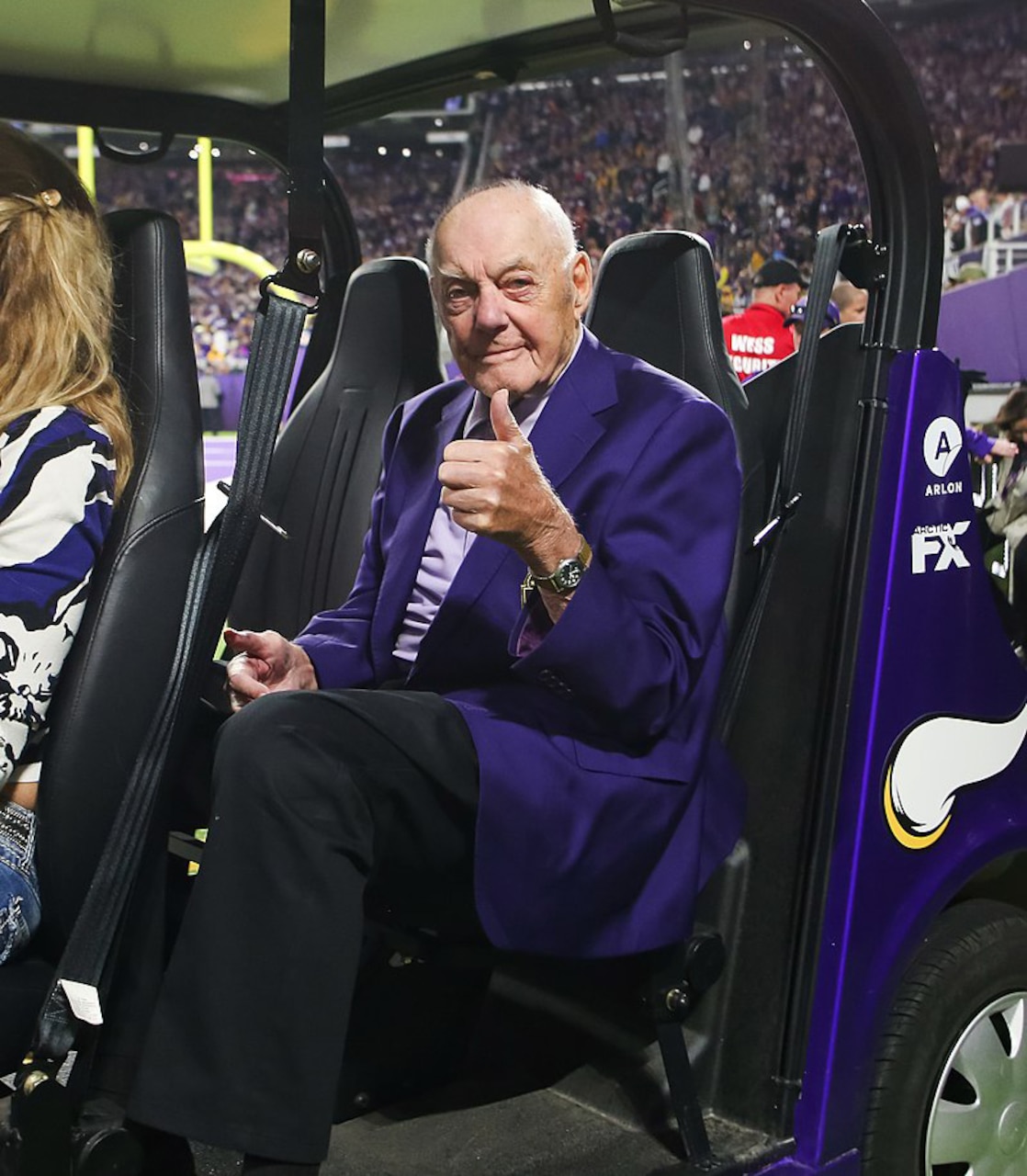 A man sits in a chair in a crowded spectator area and gives the thumbs up sign.