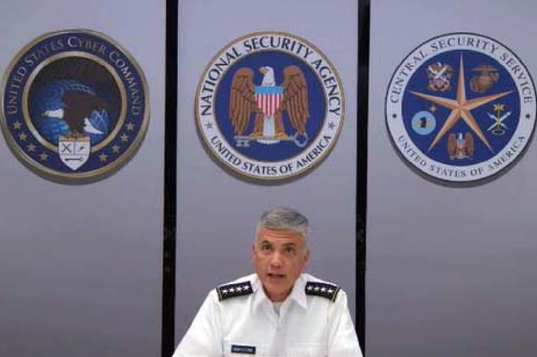 A man wearing a uniform sits and looks into a camera and speaks. Three large emblems are on the wall behind him.