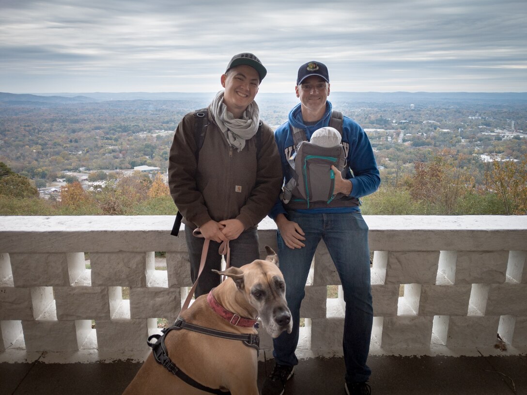 Two men, a dog, and a baby stand against a white bannister railing.
