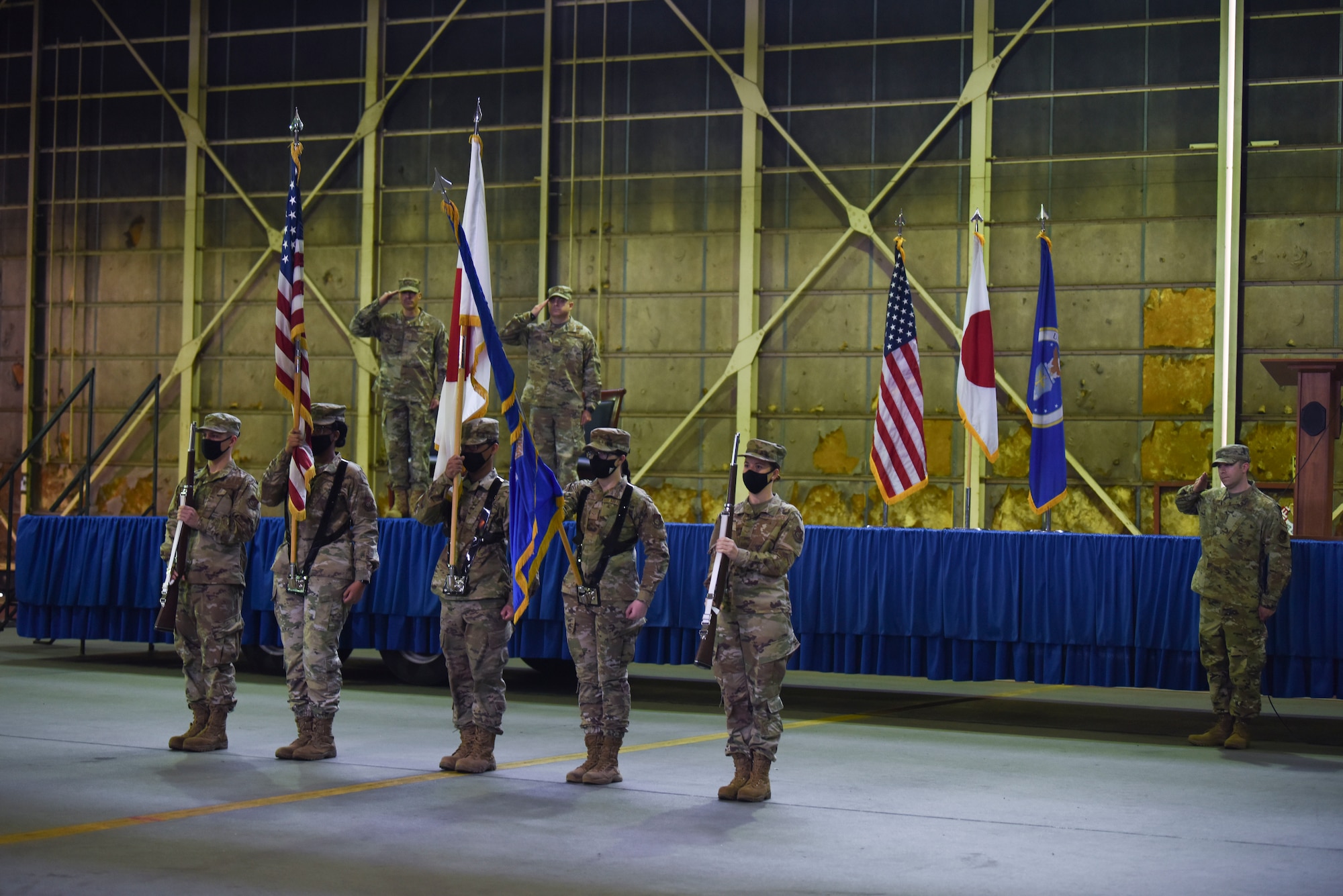 Military members hold both the U.S. flag and Japanese and rifles in front of a stage while other military members salute.