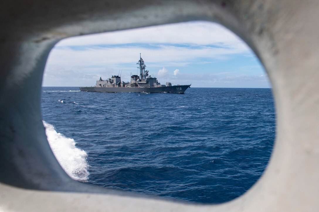 A Japanese ship sailing in the distance is framed by an oval-shaped opening on a Navy ship.