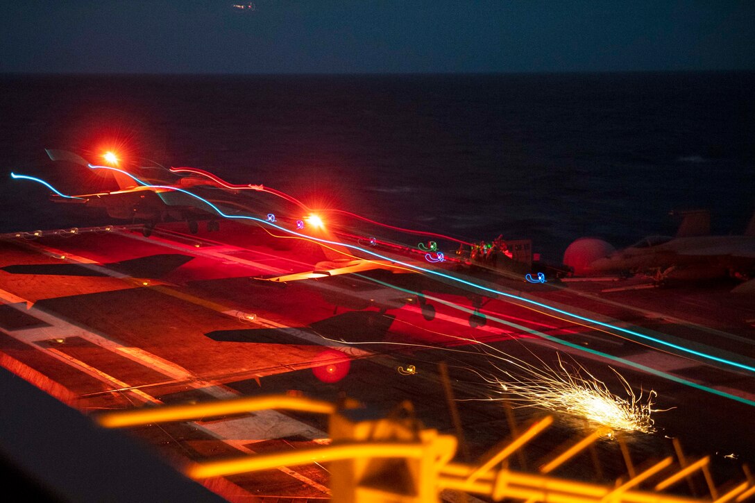 An aircraft lands on a ship at sea illuminated by colorful lights.