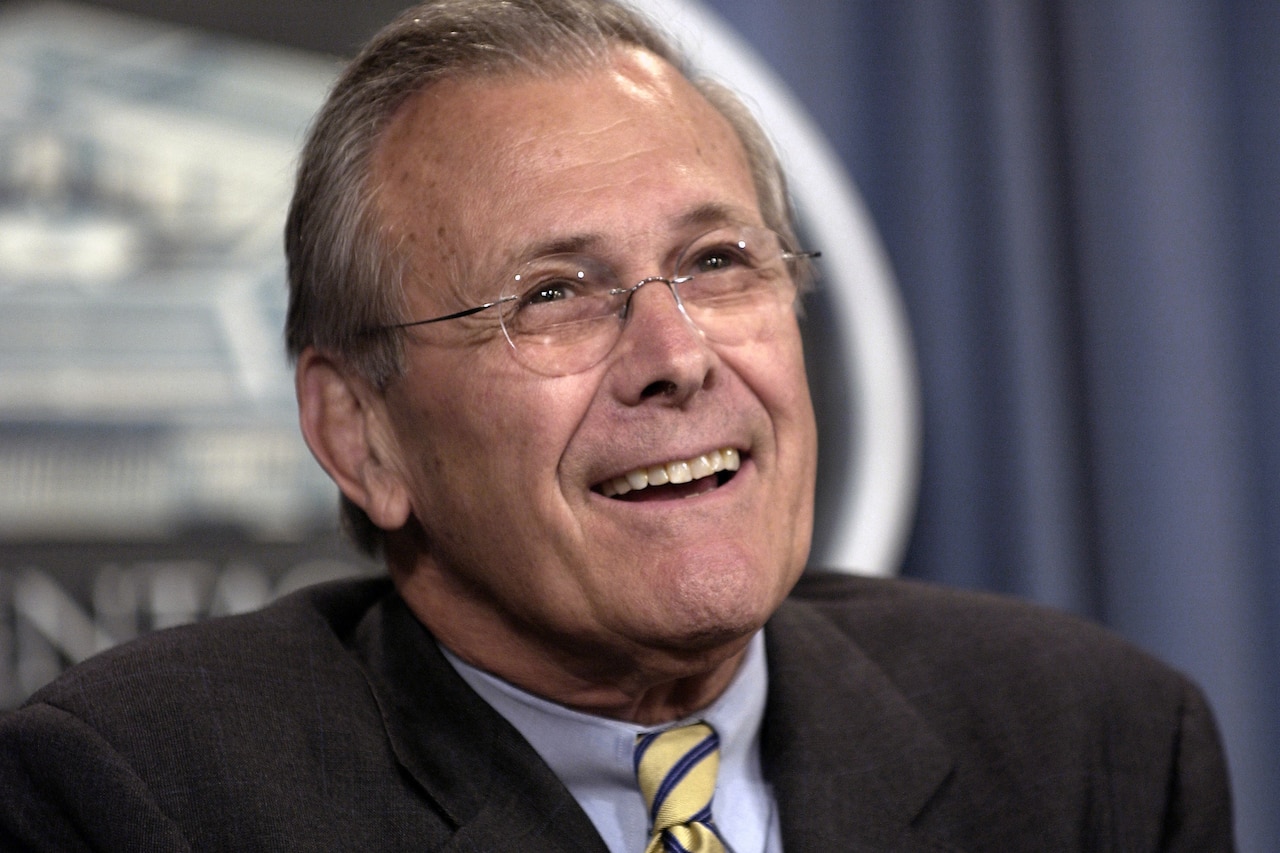 Defense Secretary Donald H. Rumsfeld smiles while sitting in front of a blue curtain and Pentagon logo.