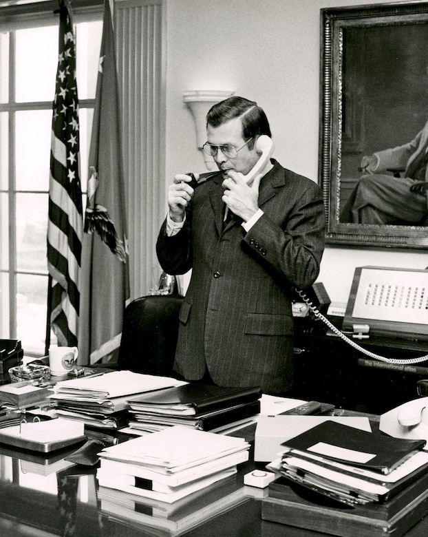Defense Secretary Donald H. Rumsfeld stands and smokes a pipe while holding a phone.