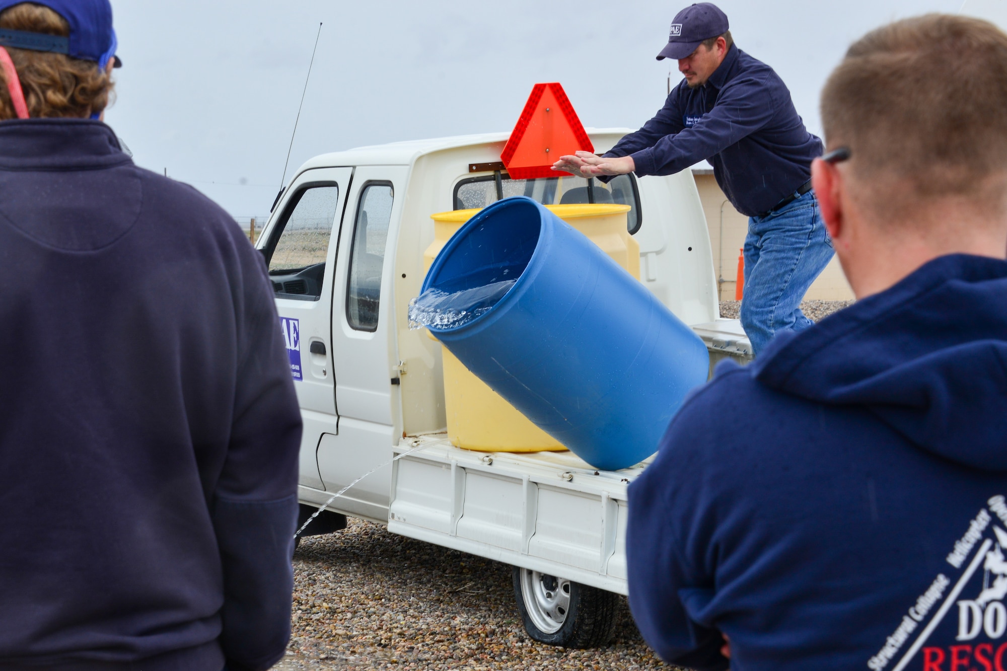 Man tips over a container of water to demonstrate what a 30 gallon spill looks like to a small group of people at Kirtland, AFB.