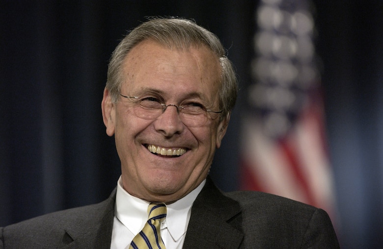 Defense Secretary Donald H. Rumsfeld smiles while standing in front of a blue curtain and U.S. flag.